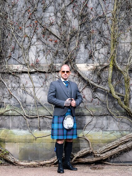 A person in traditional Scottish attire standing in front of a wall with bare branches. 4
