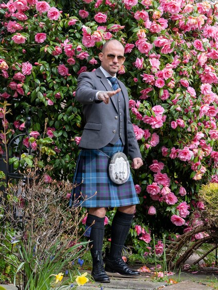 A person in a kilt and jacket standing in front of a bush of pink flowers. 2