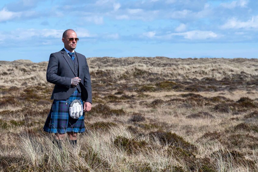 A man in a suit and kilt standing in a grassy field with a cloudy sky. 5