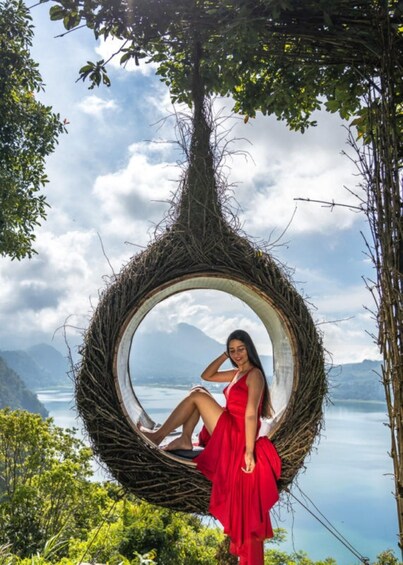A person in a red dress sitting in a circular wicker structure with a scenic lake and mountains in the background. 2