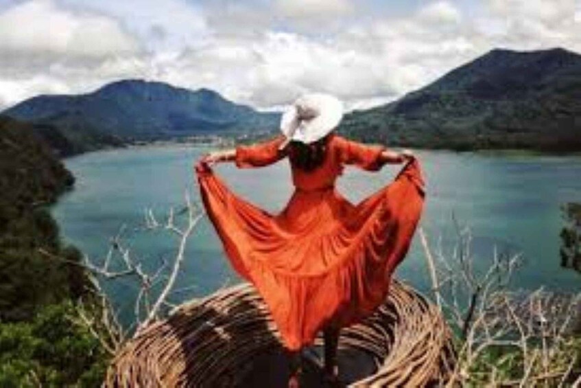 A person in a red dress standing on a woven basket by a lake with mountains in the background. 5