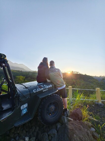 Dos personas sentadas en un vehículo con una placa de matrícula, contemplando un paisaje montañoso con el sol poniéndose al fondo. 4
