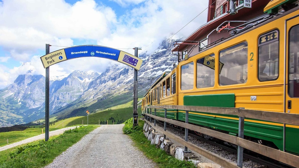 A train at a station with a mountainous backdrop, a sign for Brandegg and Grindelwald, and a path leading to the station. 4