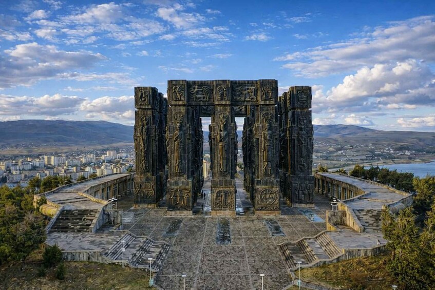 A large stone structure with intricate carvings, surrounded by a cityscape and mountains in the background. 1