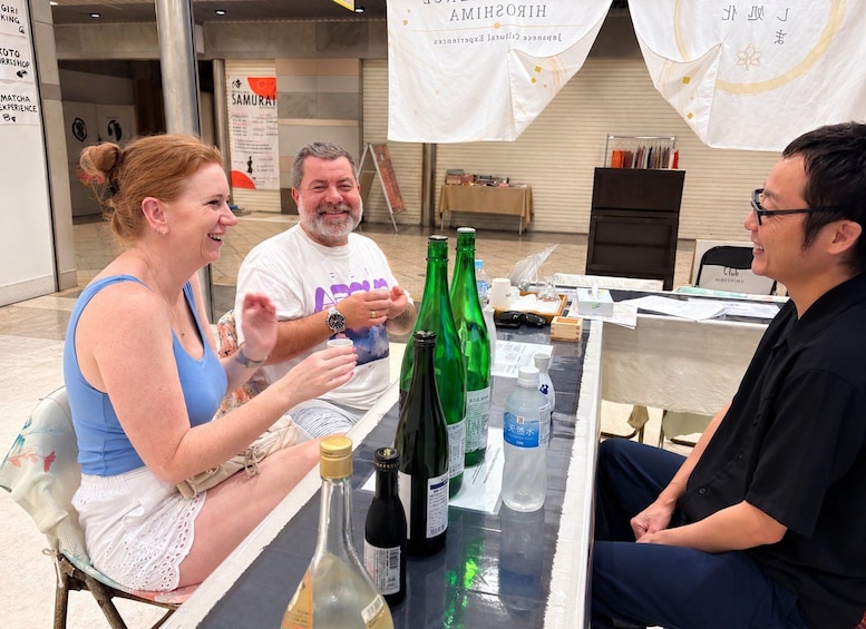 Three people seated at a table with various bottles and a laptop, in a room with banners and a poster in the background. 1
