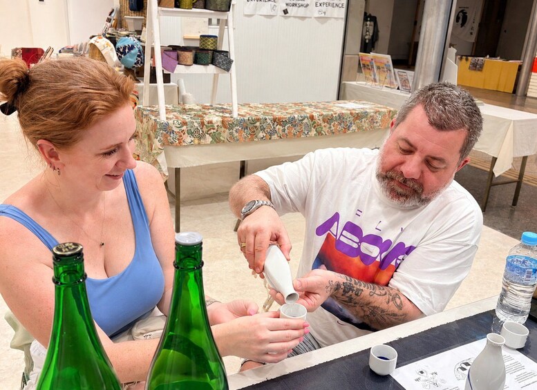 Two people at a table with a patterned tablecloth, a green bottle, and a white cup. 2
