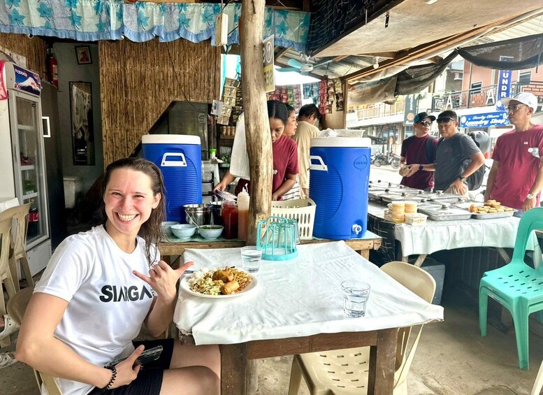 A person sitting at a table with a plate of food and a drink, in a casual outdoor setting with other people and food stalls in the background. 2