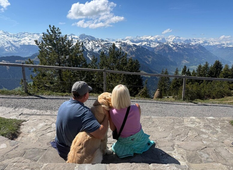 A man and a woman with a dog sitting on a stone ledge overlooking snow-capped mountains. 1