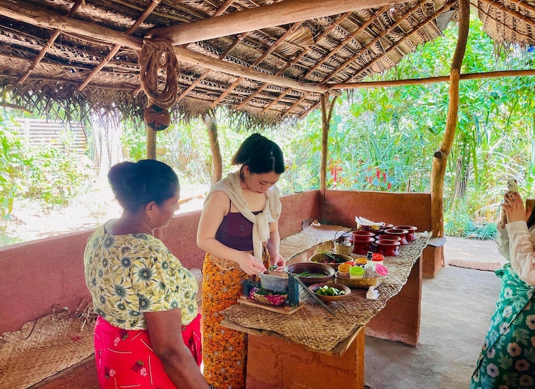 A rustic outdoor setting with a thatched roof structure, a woven table with various food items, and lush greenery in the background. 1