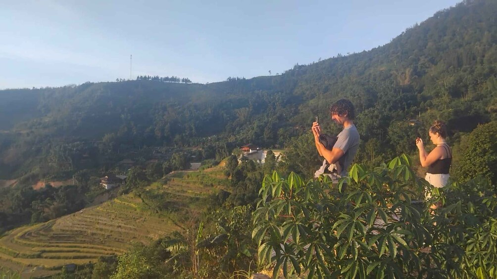 Two people meditating on a hillside overlooking terraced fields and a forested hill. 5