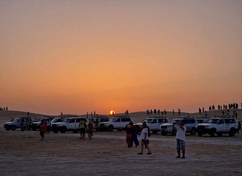 A desert landscape with a sunset, people walking, and parked vehicles. 1