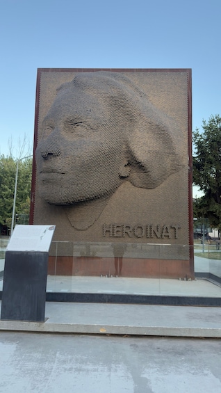 A large sculpture made of cylindrical objects, with a plaque reading 'HEROINAT' below it. 4