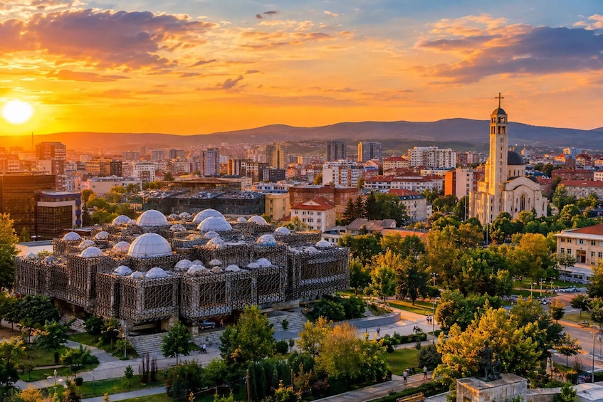 A cityscape at sunset with a prominent mosque featuring multiple domes, surrounded by modern buildings and a large church with a tall bell tower. 1
