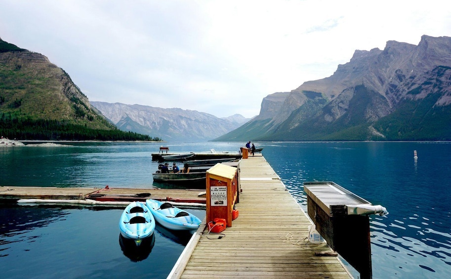 A wooden dock extending into calm water with kayaks docked, a fishing sign, and mountains in the background. 3