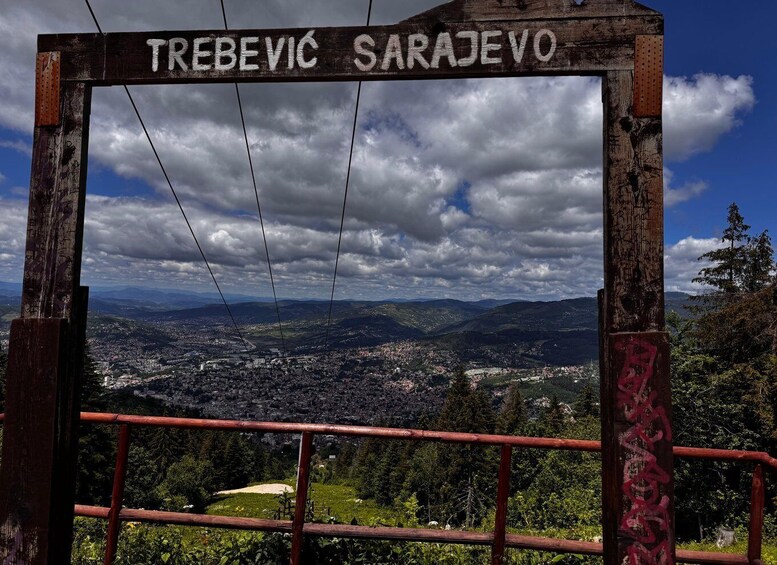 A wooden sign with 'TREBEVIC SARAEVO' written on it, overlooking a town and mountains. 3