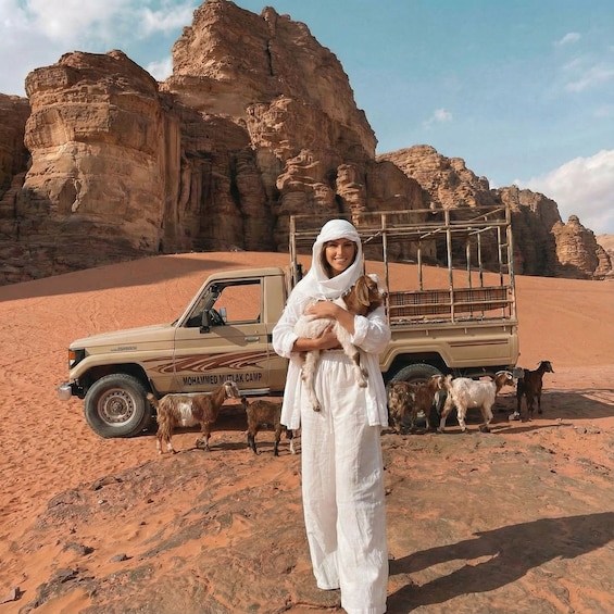 A person in white attire holding a goat, with a beige off-road vehicle labeled "MOHAMMED MTLAK CAMP" and several goats in the background. 1
