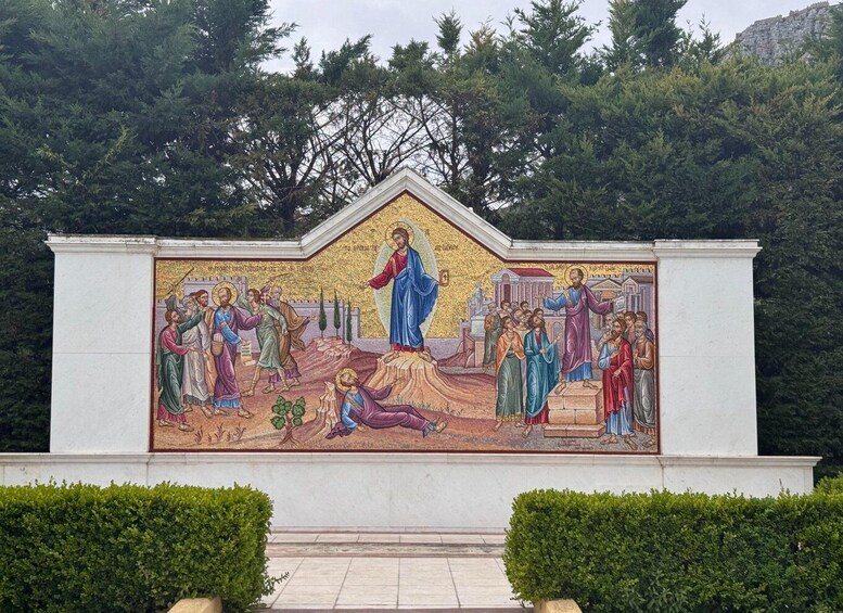 A mosaic mural on a white wall with a central figure, surrounded by smaller figures, set against a backdrop of trees and a hill. 1