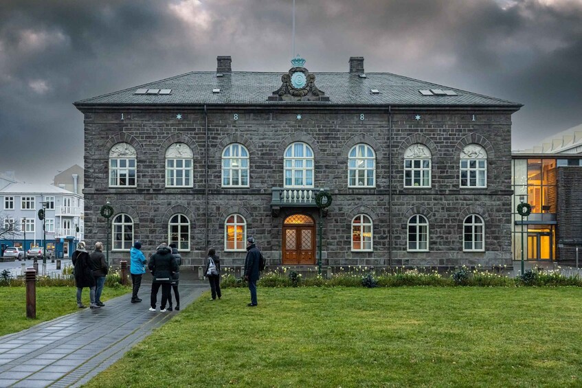 A historic stone building with arched windows and a clock tower, surrounded by a grassy area and a few people. 1