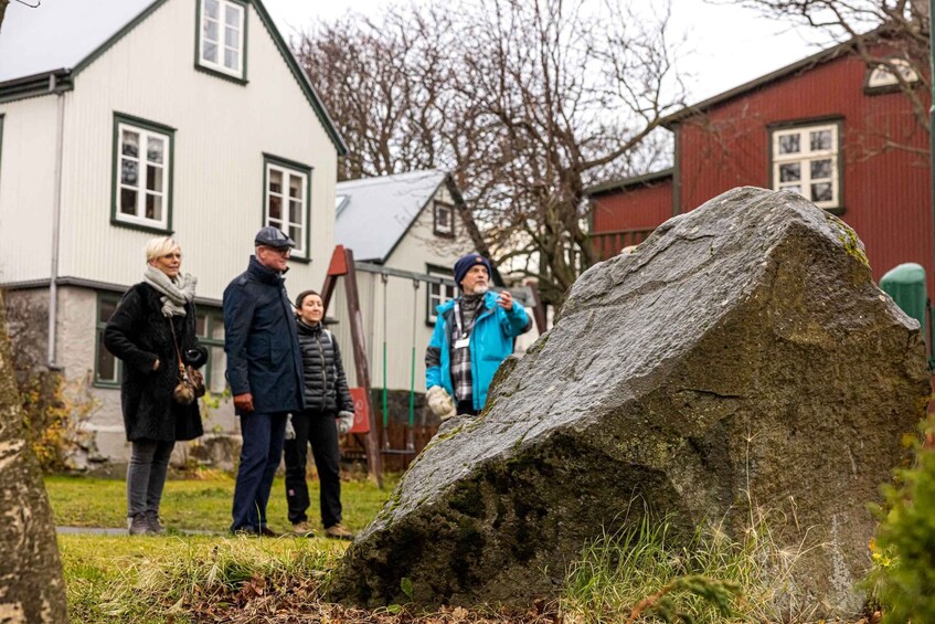 A group of people standing around a large rock in a grassy area with houses in the background. 3