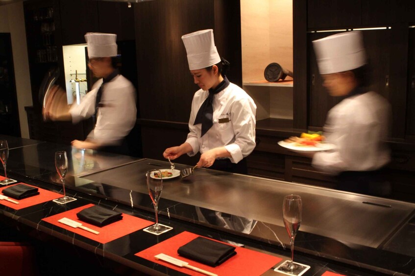 Chefs preparing food in a professional kitchen with a long counter and wine glasses. 1