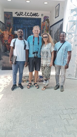 Four people standing in front of a building with a 'Welcome' sign, a colorful mural, and a decorative door. 2