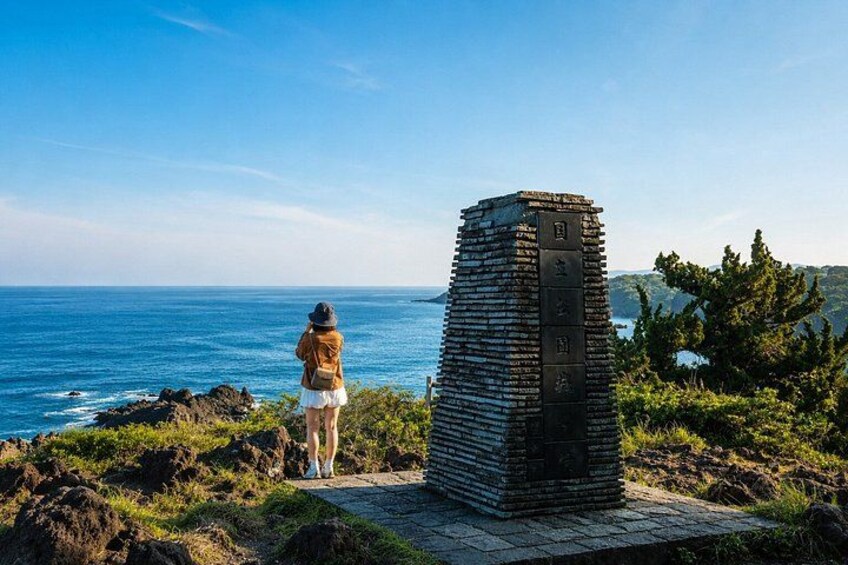 A person standing on a rocky outcrop overlooking the ocean, with a stone monument in the foreground. 2