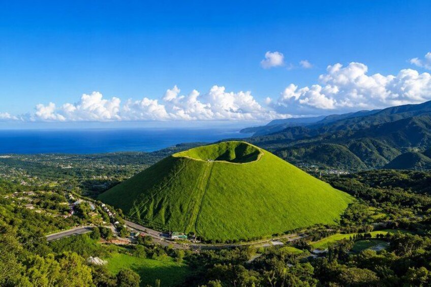 A green hill with a road and buildings in the foreground, and a coastline in the background. 1