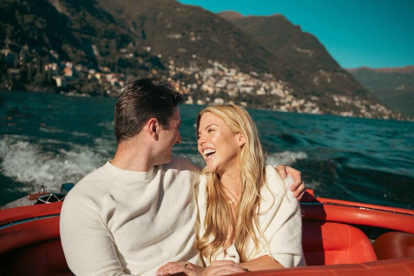 A couple on a red boat with a mountainous coastline in the background. 1
