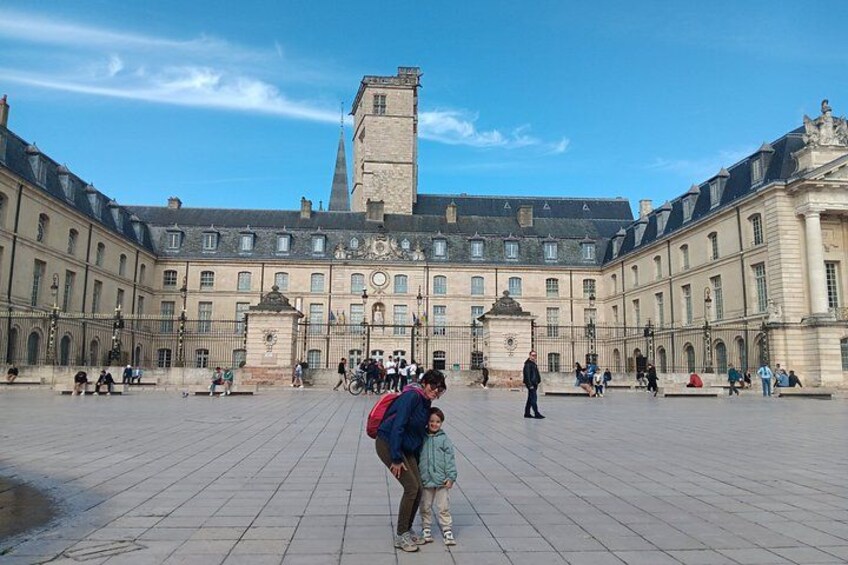 A historic courtyard with a large building featuring a tower, a fountain, and people walking. 1