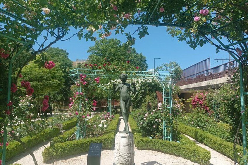 A statue in a garden with a metal archway, surrounded by various flowering plants and a pathway. 2