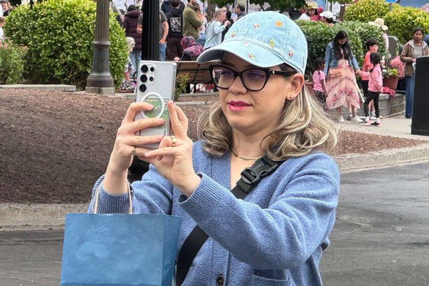 A person taking a photo with a smartphone, holding a blue shopping bag, and wearing a light blue cap and sweater. 4