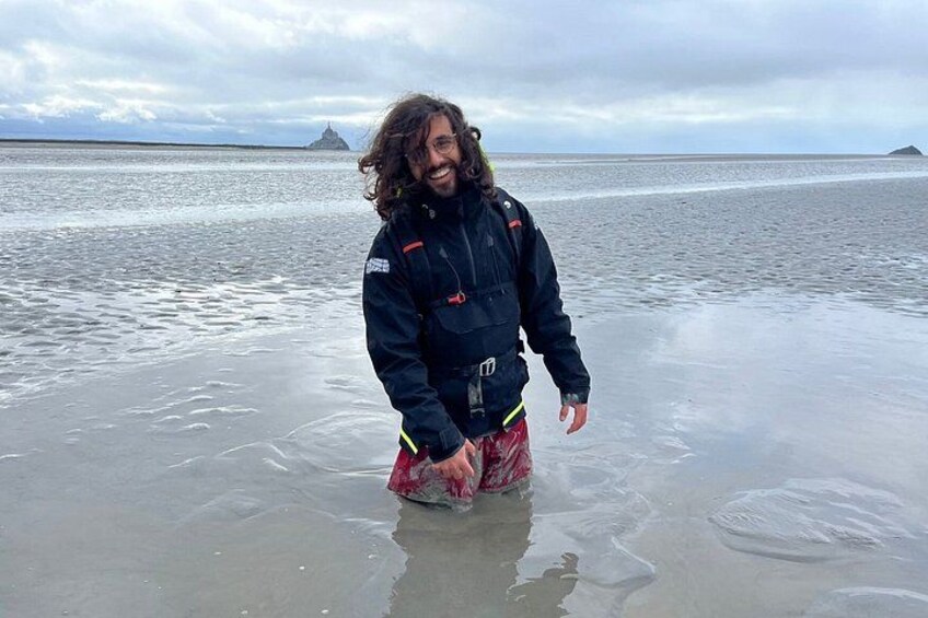 A person standing in shallow water on a beach with a cloudy sky and distant landforms. 1