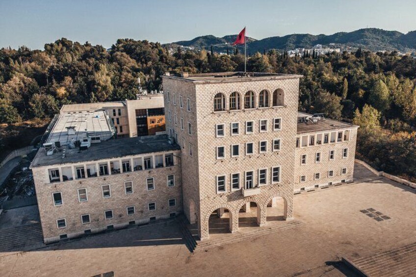 A multi-story building with a red flag on top, surrounded by trees and hills. 1