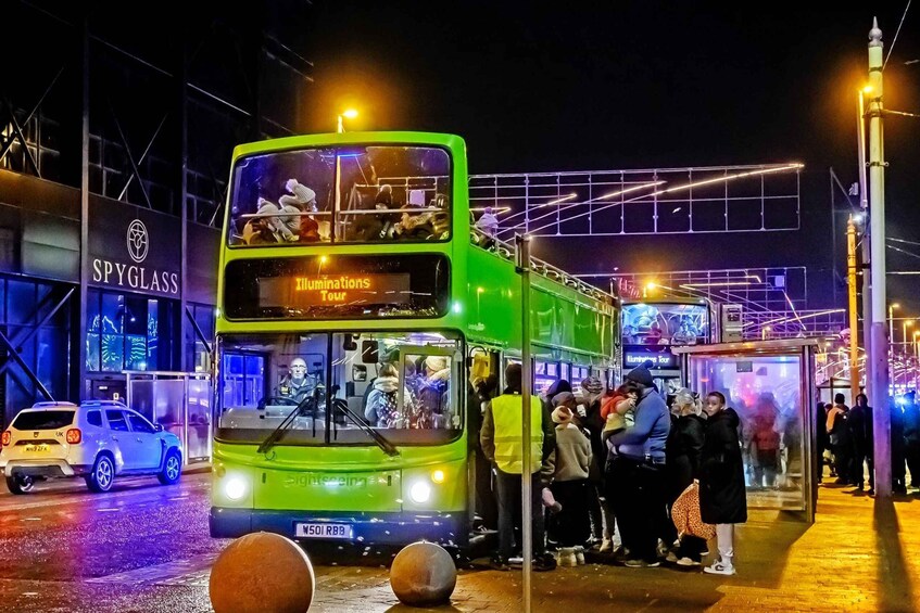 A green double-decker bus with 'Illuminations Tour' written on the front, parked at a bus stop with people waiting. 4