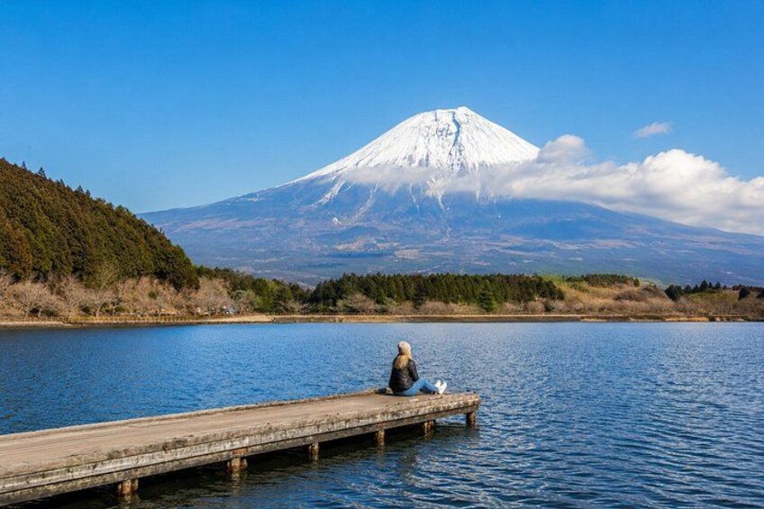 A person sitting on a wooden dock extending into a lake, with a snow-capped mountain in the background. 2