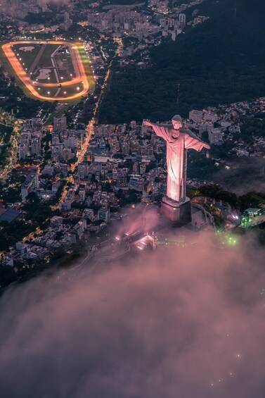 A nighttime aerial view of a city with a prominent statue illuminated by pink lights, surrounded by dense urban buildings and a lit sports field. 4