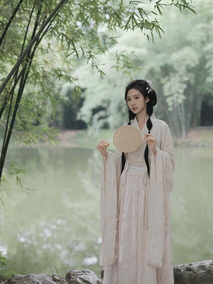 A person in traditional attire holding a fan, standing by a calm body of water surrounded by bamboo trees. 2