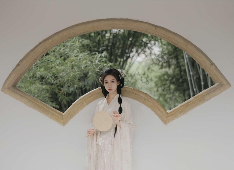 A person in traditional attire holding a fan, standing in front of a wooden-framed mirror with a bamboo background. 1