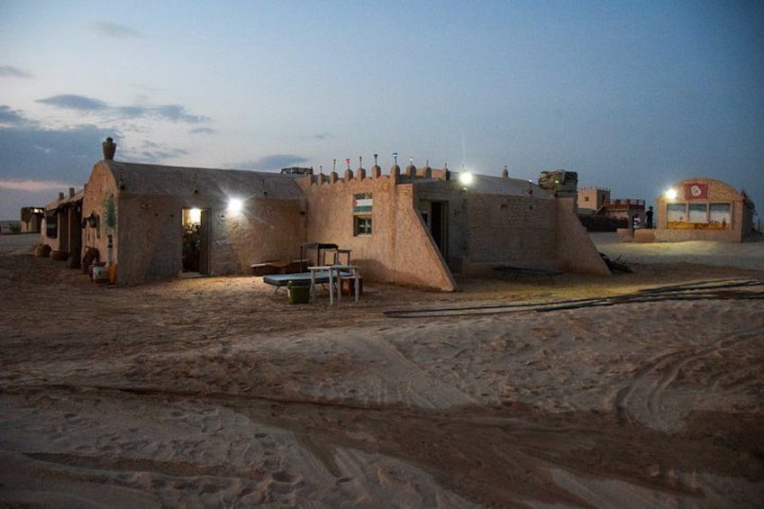Un edificio con un estilo arquitectónico singular, un espacio pavimentado y una estructura distante con luces. 1