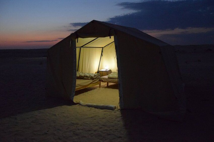 Una carpa con una cama y una lámpara en su interior, ubicada en una playa arenosa al atardecer. 3