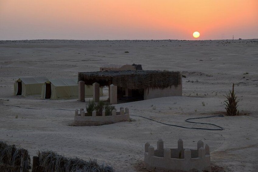 Paisaje desértico con un atardecer, tiendas de campaña, una estructura de paja y un pequeño jardín. 5