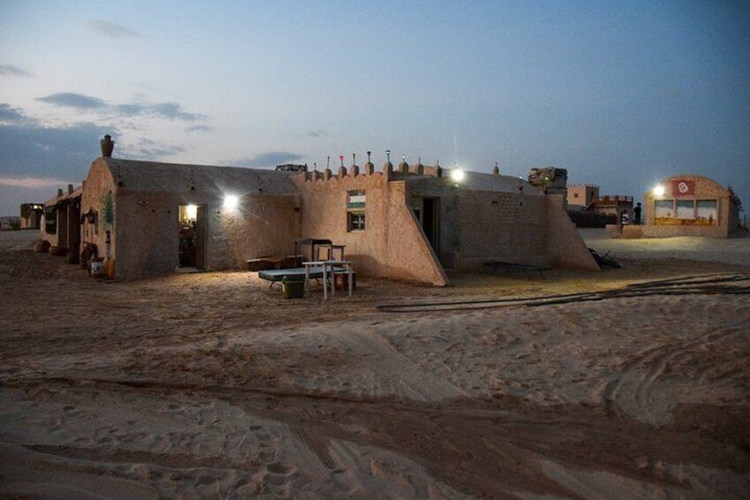A desert landscape with a small building with a flat roof and a chimney, surrounded by sand. 5