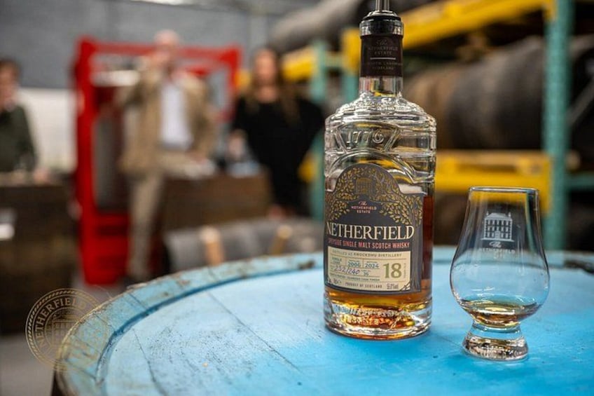 A bottle of Netherfield whisky and a glass on a blue barrel. 1