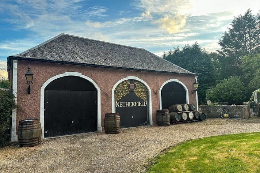 A brick building with three arched doors, barrels stacked outside, and a gravel driveway. 2