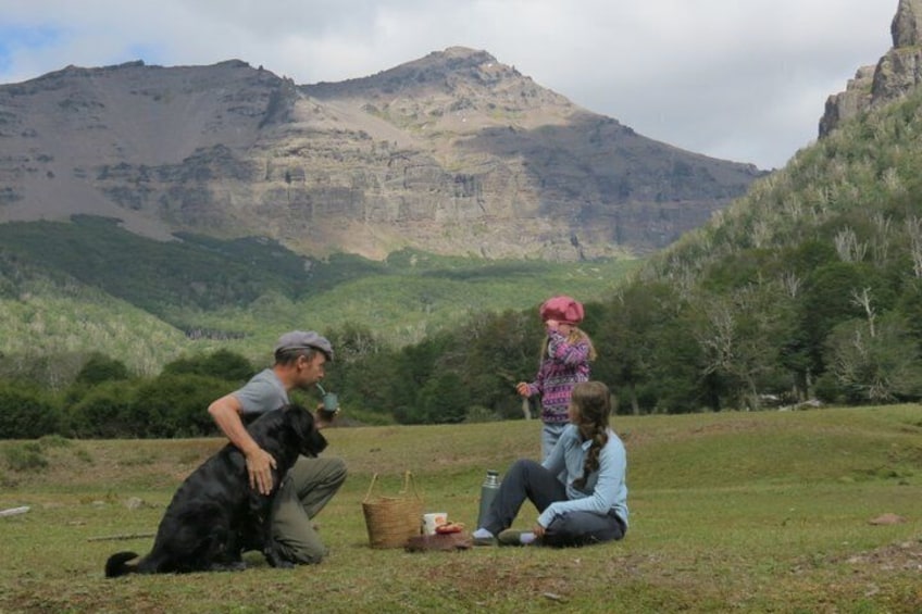 A person sitting on the grass with a dog, another person standing, and a mountain in the background. 1