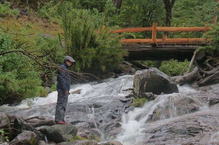 A person fishing by a flowing river with a wooden bridge in the background. 2