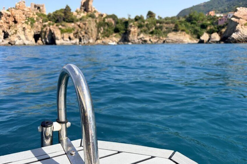 A boat with a metal railing, calm blue water, and rocky coastline with buildings. 1