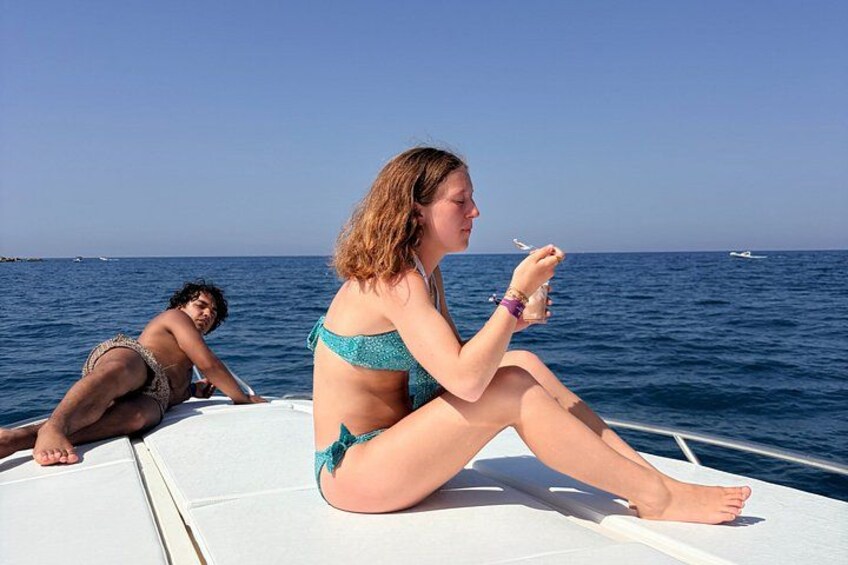 Two people on a boat, one sitting and one lying down, with the ocean and a clear sky in the background. 2