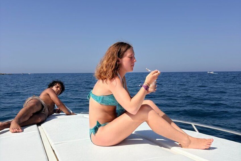 Two people on a boat, one sitting and one lying down, with the ocean and a distant boat in the background. 2