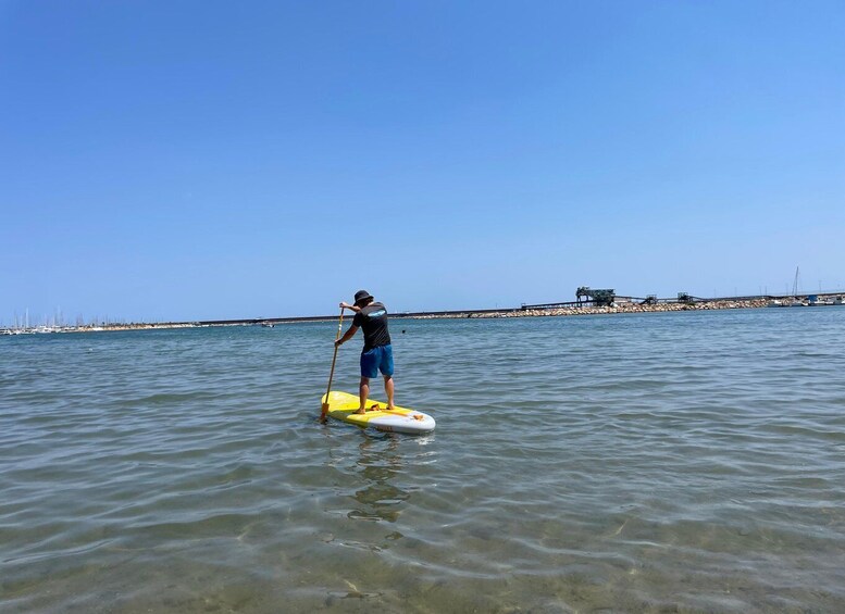 A person paddleboarding on clear water with a distant shoreline and structures. 3